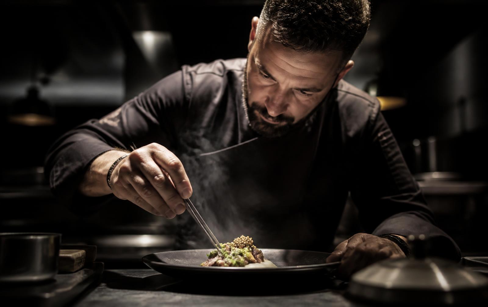 A cook finishing a plate with tweezers in a dark professional kitchen