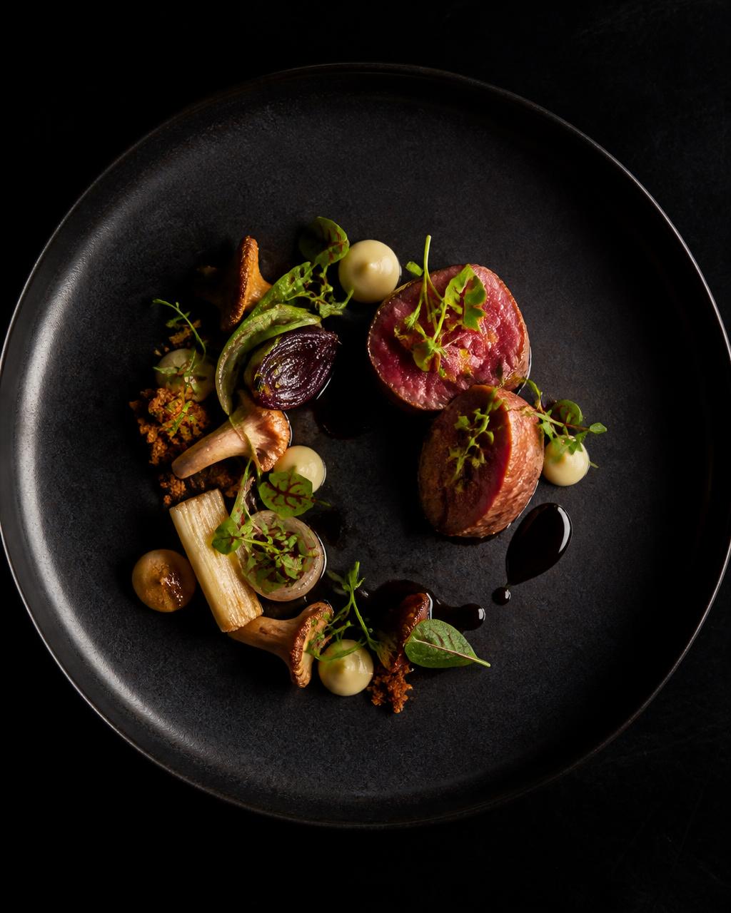Precisely plated dish of venison, mushrooms and micro herbs on matte black ceramic